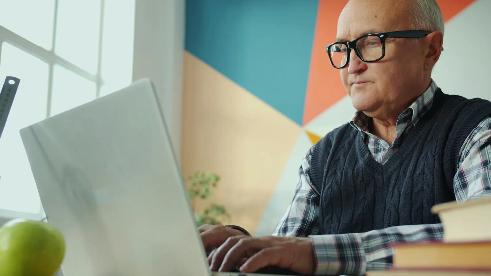 Un homme d'une soixantaine d'années, lunettes et gilet sans manches, travaille sur son ordinateur portable dans un bureau coloré.