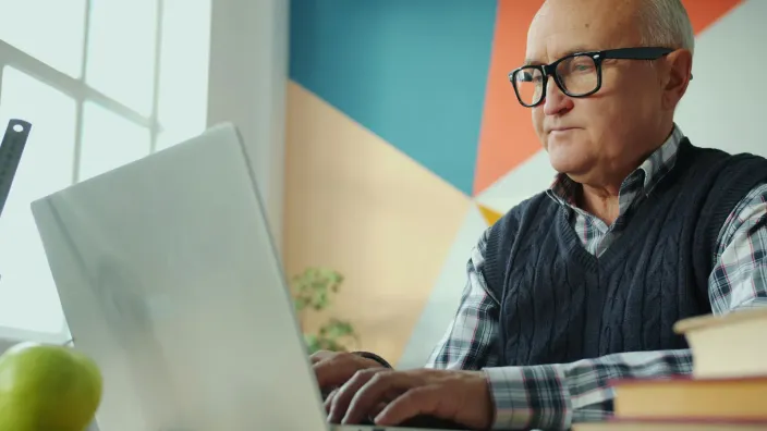 Un homme d'une soixantaine d'années, lunettes et gilet sans manches, travaille sur son ordinateur portable dans un bureau coloré.